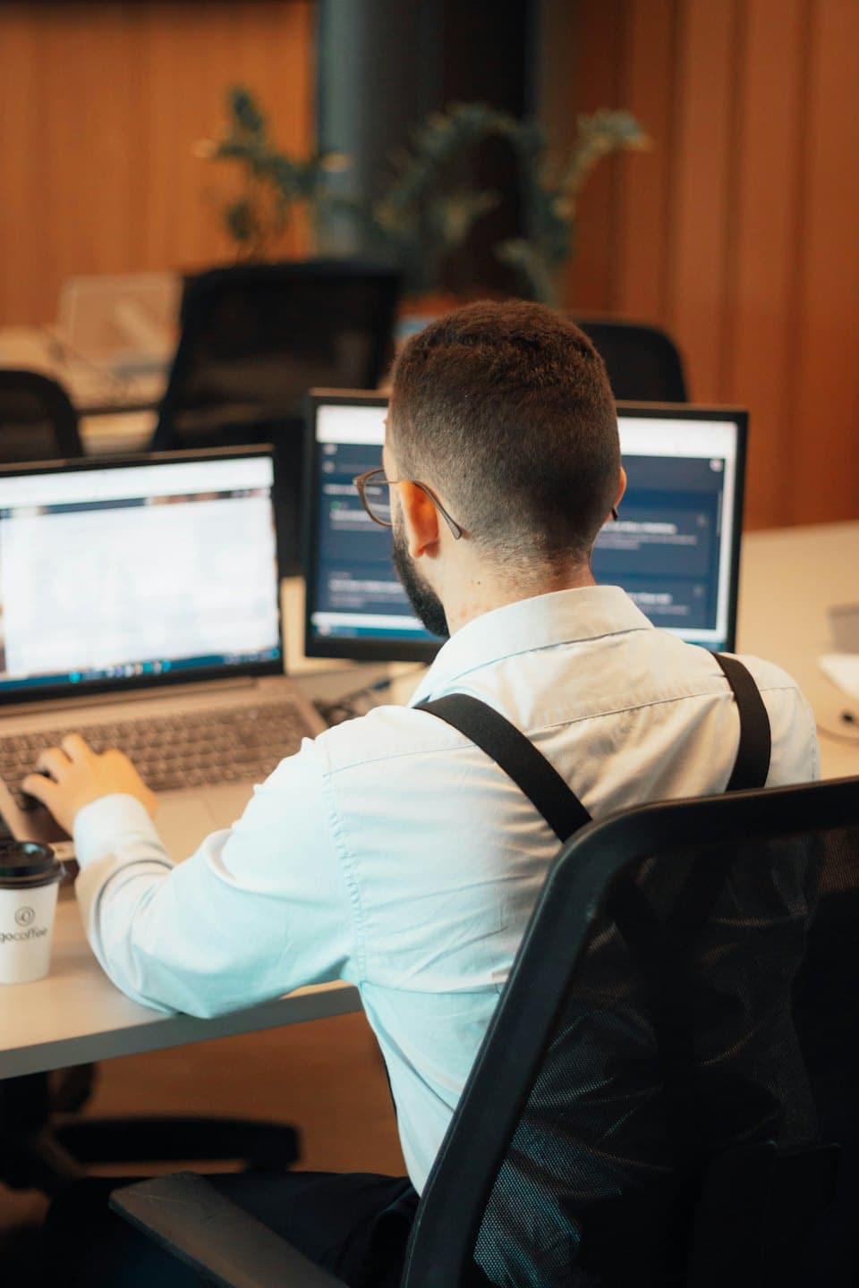 Man in suspenders works on laptop and monitor at desk.